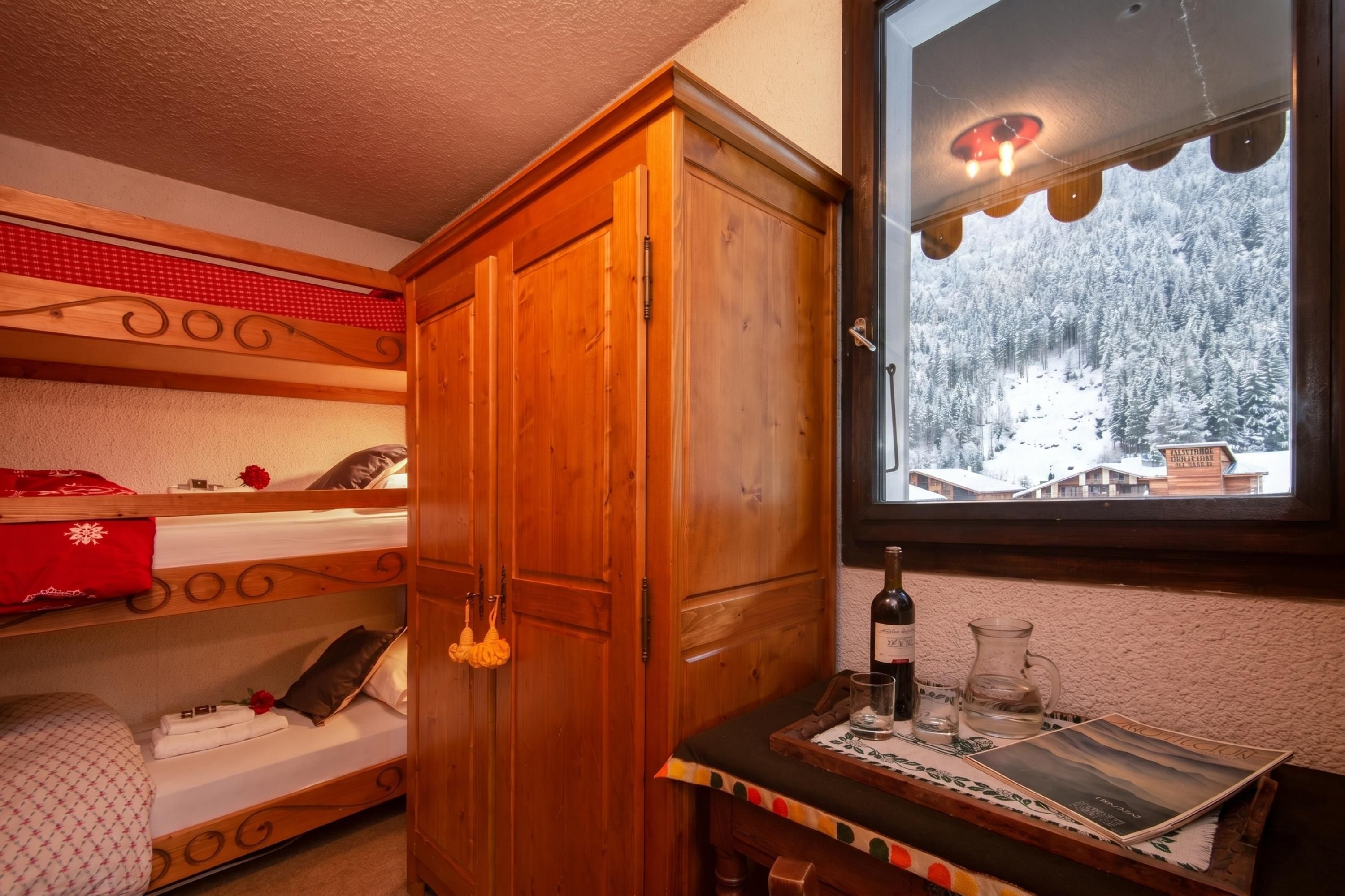 Une chambre avec un lit superposé, une armoire en bois, une fenêtre donnant sur un paysage enneigé et une table avec une bouteille et des verres.