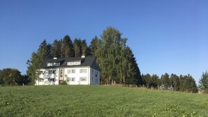 Una casa de dos pisos con techo inclinado, rodeada por un campo cubierto de césped y árboles frondosos.