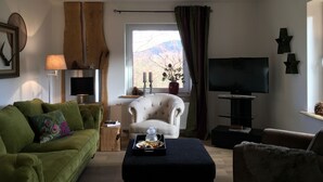 Una sala de estar con un sofá verde, un sillón blanco, un reposapiés negro, un televisor y una ventana con vistas a montañas.
