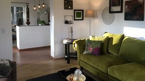 Una sala de estar con un sofá verde, una mesita auxiliar con una vela y una lámpara. Hay cuadros en la pared y una ventana con vistas a árboles.