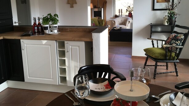 Una cocina moderna con una isla central, armarios blancos y un comedor con una mesa y una silla negra dispuestas para dos.
