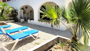 Um pátio com cadeiras de lounge azuis, um edifício branco com arcos e plantas em vasos.