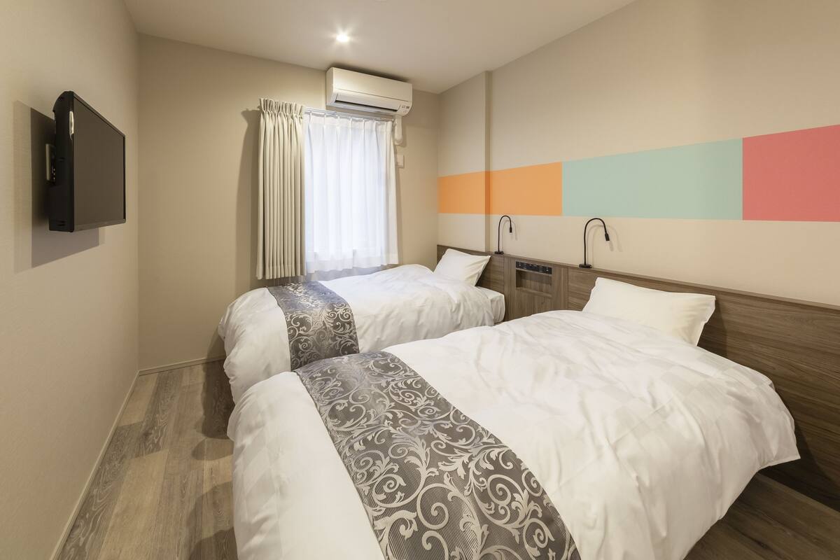 A hotel room with two beds, an air conditioning unit, a flat-screen TV, and a colorful wall design.
