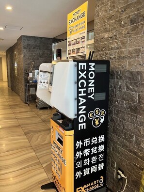 A money exchange machine with instructions in multiple languages, located in a hallway with a stone wall and a wooden floor. - SARASA HOTEL Dotonbori (Osaka)