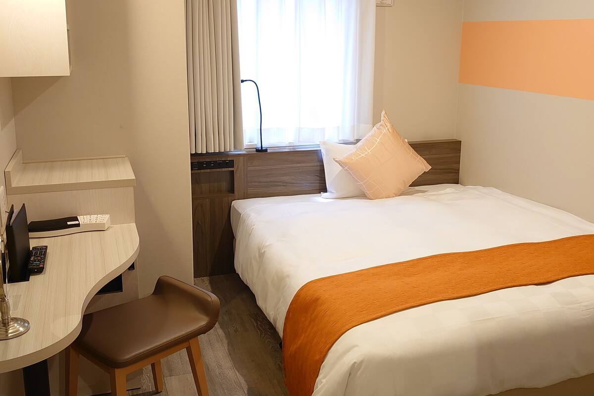 A hotel room with a bed, a desk with a chair, a window with curtains, an air conditioning unit, and a bedside table.