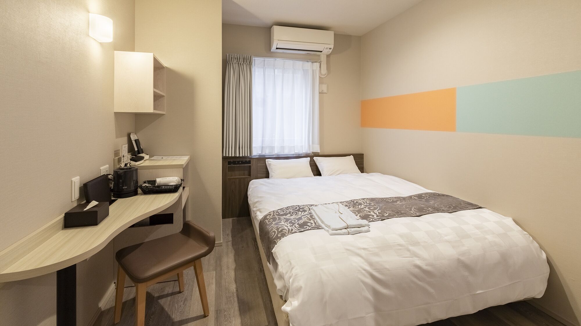 Ett hotellrum med en säng, ett skrivbord, en stol, en luftkonditionering och ett fönster med gardiner.
