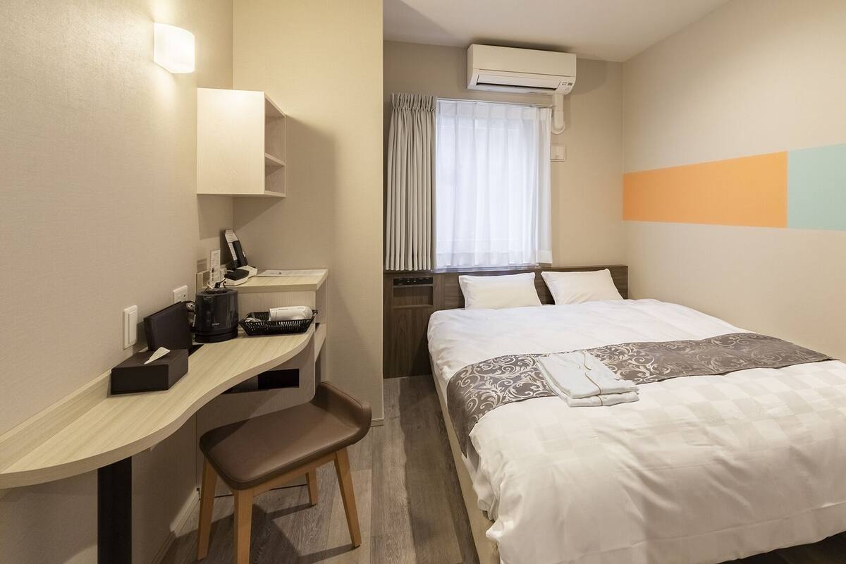 A hotel room with a bed, a desk, a chair, an air conditioner, and a window with curtains.