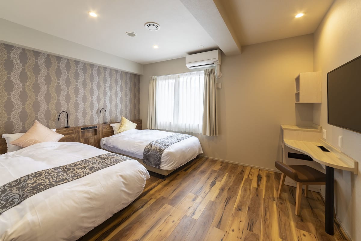 A hotel room with two beds, a wooden floor, a desk with a chair, an air conditioning unit, and a window with curtains.