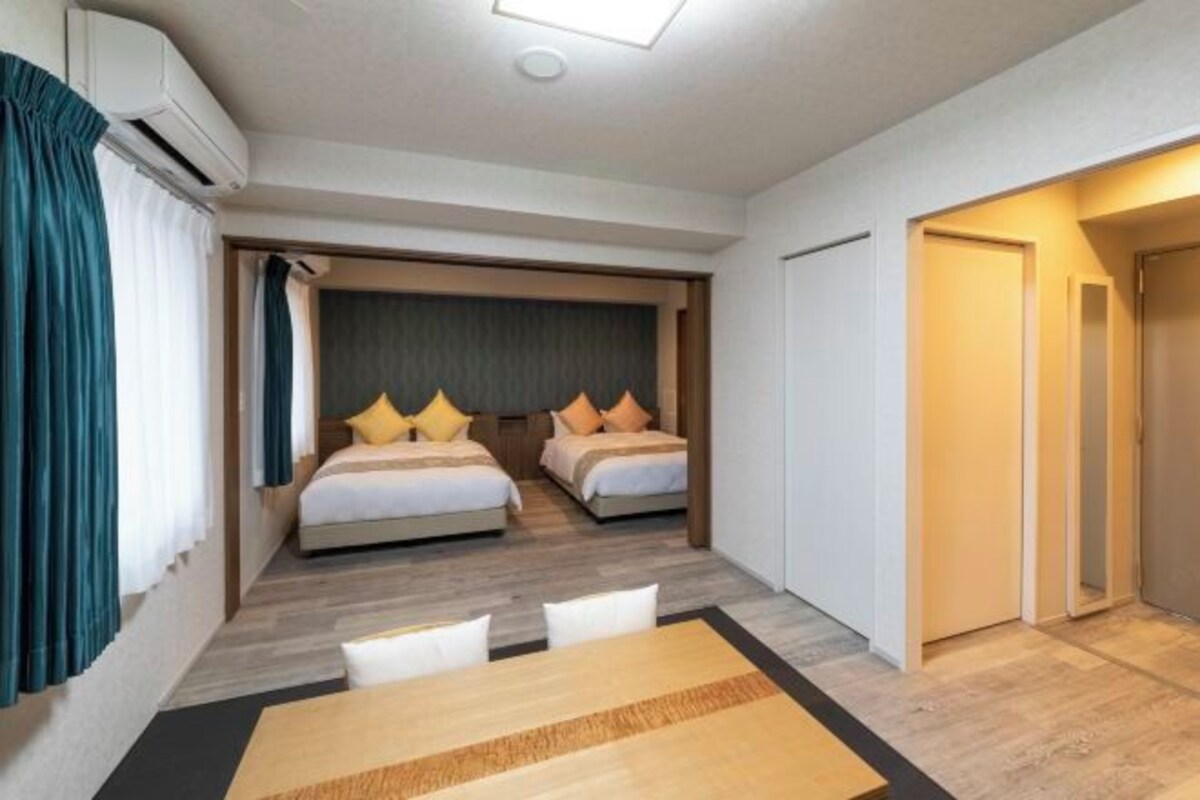 A modern hotel room with two beds, a dining table, and a separate door leading to the bathroom.