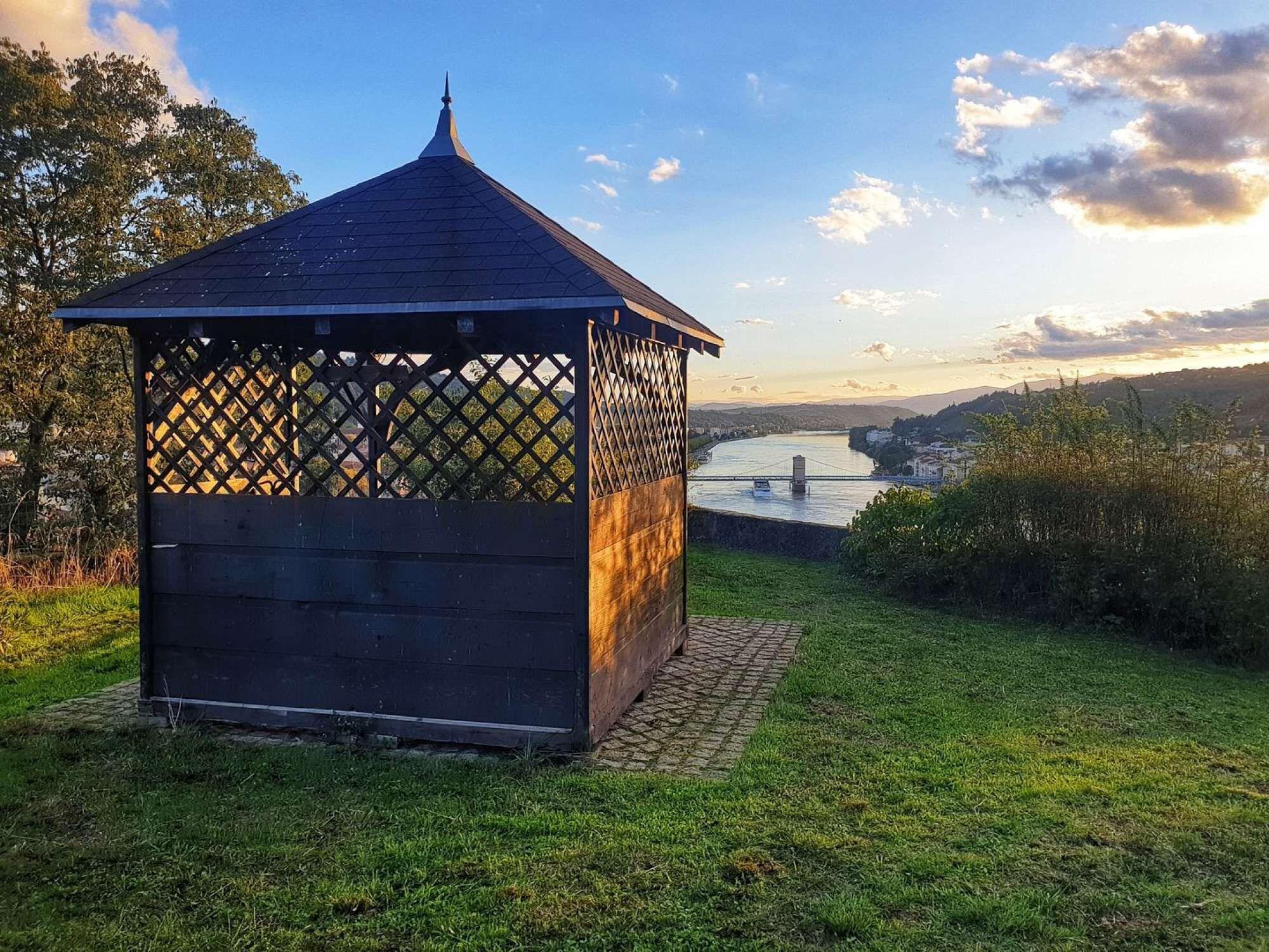Ein Gazebo mit Gitterdach, eingebettet in eine grüne Wiese, im Hintergrund ein Fluss und eine Brücke.