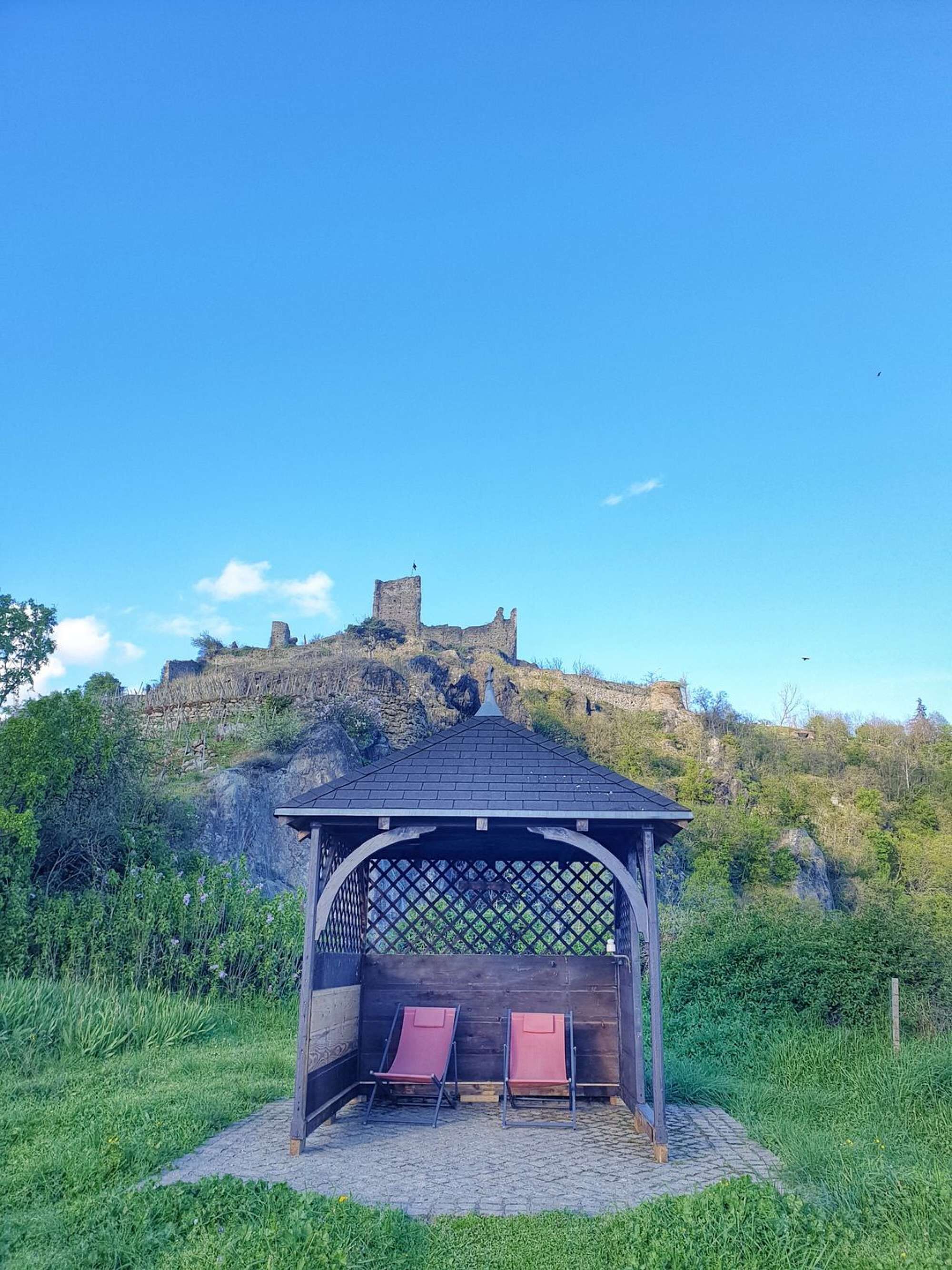 Ein Gartenpavillon mit zwei roten Stühlen und einem Gitter an der Vorderseite, gelegen auf einem Kopfsteinpflasterweg mit Blick auf einen historischen Wehrturm auf einem Hügel.