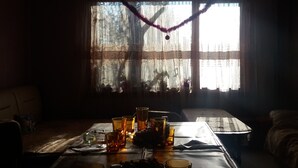 Une pièce faiblement éclairée, avec un plafond en bois, une table dressée avec des verres et une bouteille, et une fenêtre ornée de rideaux.