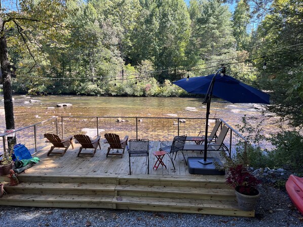 A wooden deck with outdoor seating, overlooking a river, surrounded by trees.