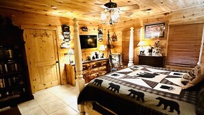 A cozy room with wooden walls and ceiling, a large bed with a bear-themed quilt, a television, and various wall decorations. - Blue Ridge Castaway Cabin on The Toccoa River (Blue Ridge)