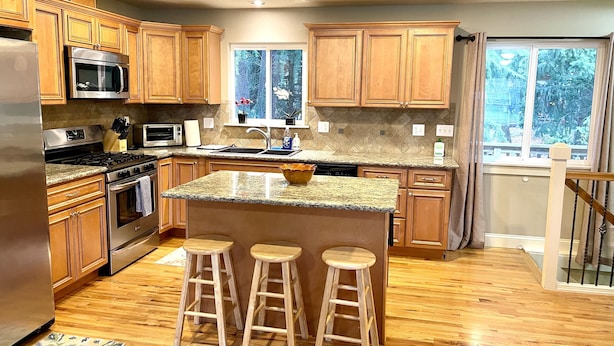 A kitchen with wooden cabinets, a granite countertop, a stainless steel refrigerator, a microwave, a stove, and three wooden stools. - Great location for visiting the NW. Comfortable 5 bedroom home. (Portland)