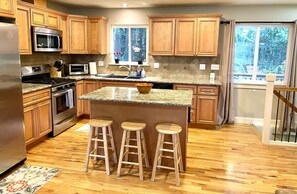 A kitchen with wooden cabinets, a granite countertop, a stainless steel refrigerator, a microwave, a stove, and three wooden stools.