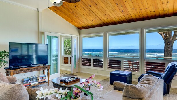 A spacious living room with a large TV, comfortable seating, a coffee table, and a view of the ocean through large windows. - Haena Beachfront With Stunning Panoramic Ocean Views TVNC # 5134  (Hanalei, Haena)