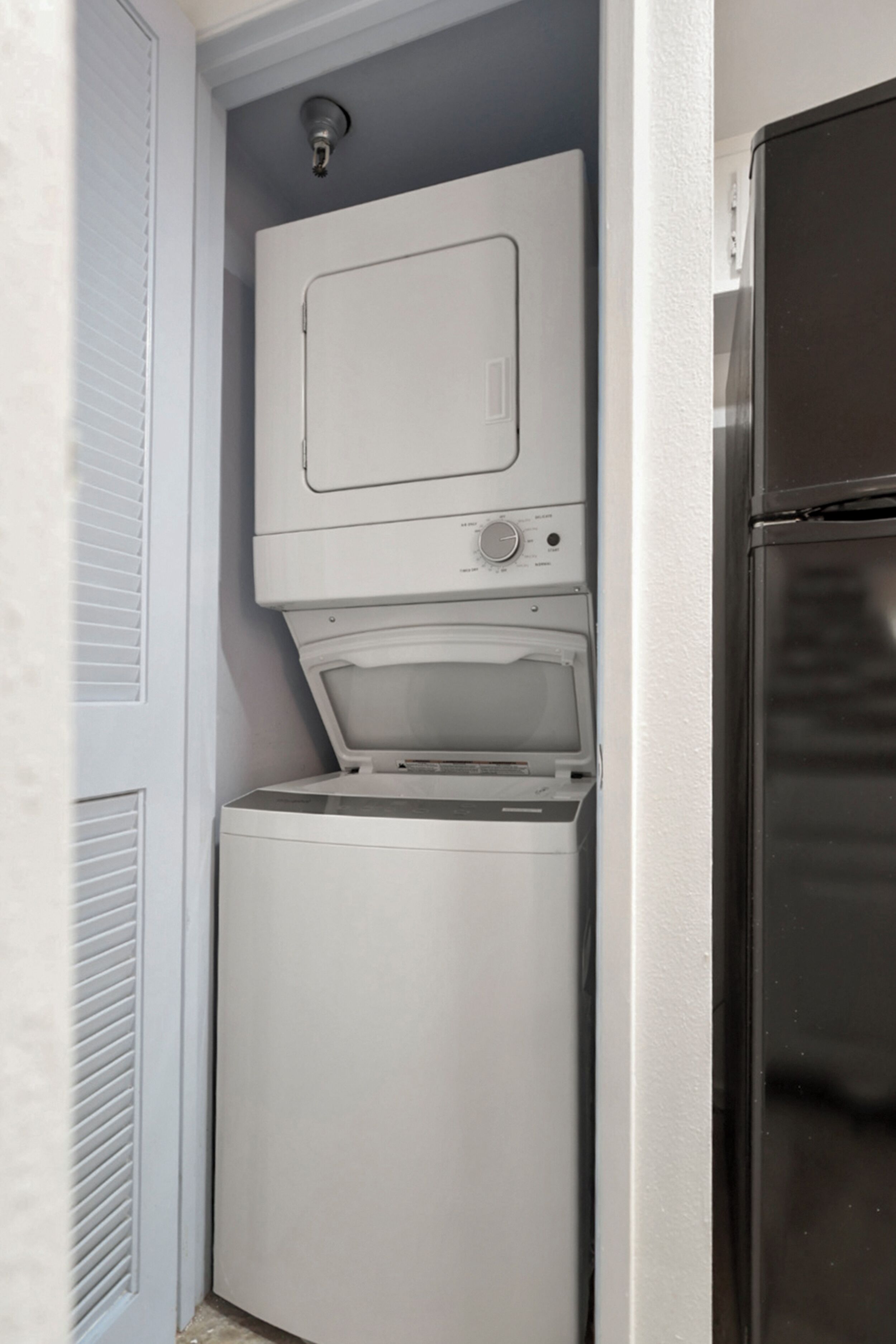 A compact laundry room with a washer and dryer stacked vertically, a refrigerator to the right, and a door with a vent on the left.