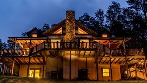A large, two-story log cabin with a stone chimney, wooden balconies, and a covered front porch. - The Lodge: Sleeps 23 - Hot Tub, Fire Pit, Game Room (Logan)