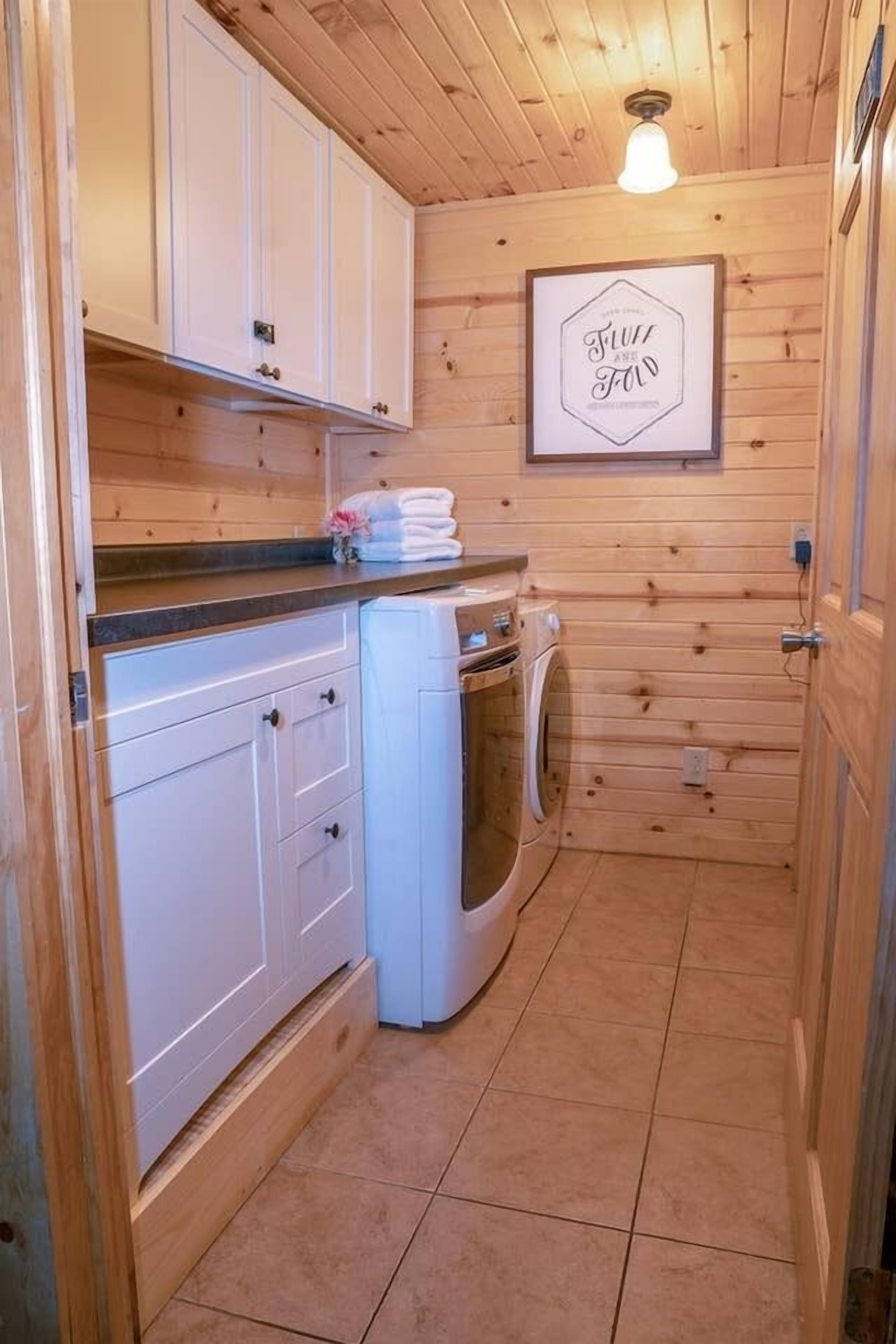 A compact kitchen with wooden walls, white cabinets, a washing machine, and a framed sign on the wall.