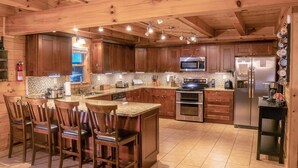 A spacious kitchen with wooden cabinets, a central island with bar stools, stainless steel appliances, and a tiled backsplash. - The Lodge: Sleeps 23 - Hot Tub, Fire Pit, Game Room (Logan)