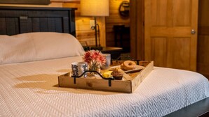 A neatly made bed with a tray of breakfast items including a muffin, a donut, and a cup, set against a wooden wall with a lamp and a painting. - The Lodge: Sleeps 23 - Hot Tub, Fire Pit, Game Room (Logan)