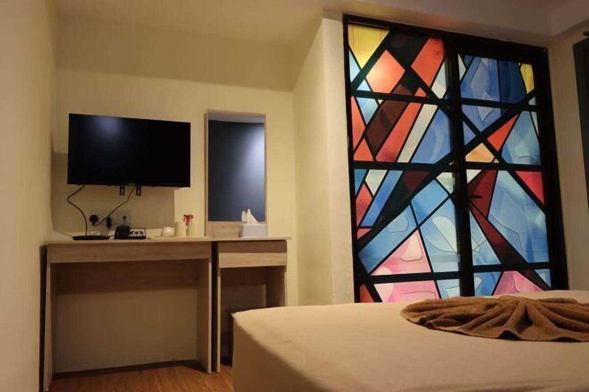 A hotel room with a bed, a desk with a flat-screen TV, a mirror, and a stained glass window.