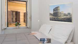 A hotel room with a bed, a window with shutters, a potted plant, a framed picture of the Colosseum, and a bedside table with a book. - Rental In Rome Arenula Apartment (Rome)