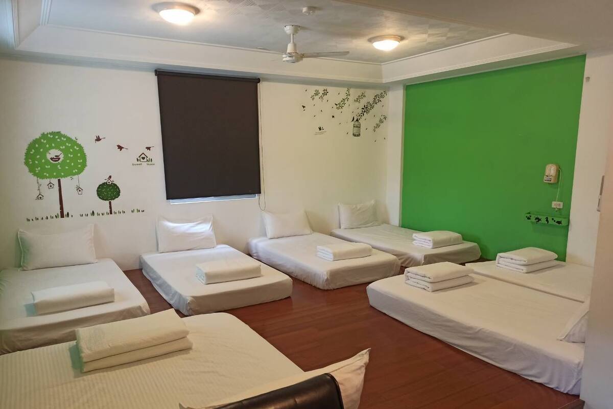 Habitació d'hotel amb quatre llits individuals, una paret d'accent verd, un ventilador de sostre i una finestra amb una persiana.