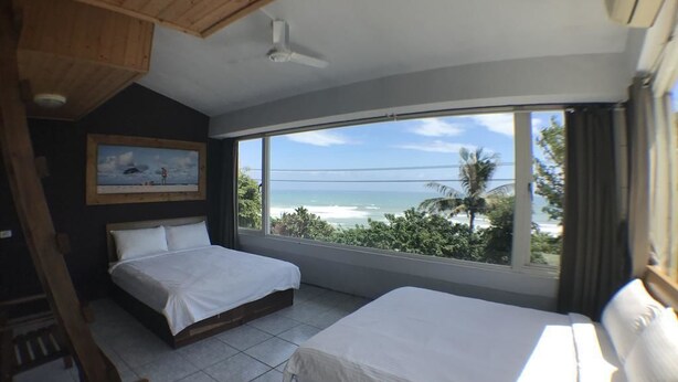 Une chambre avec deux lits, une grande fenêtre donnant sur la plage et un ventilateur de plafond.