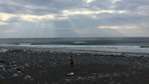 Une plage avec une personne debout sur le sable, une planche de surf et l’océan en arrière-plan.