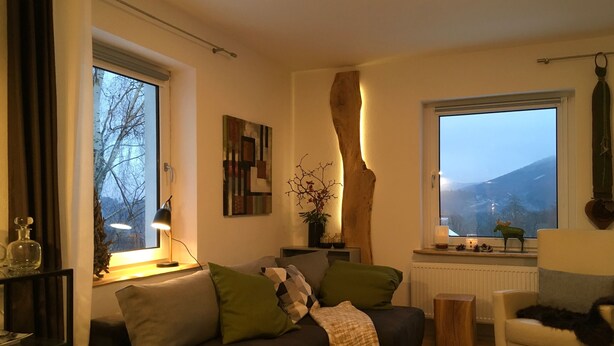 Un salón acogedor con un sofá de color oscuro, una pequeña mesa auxiliar de madera, un sillón blanco y un ventanal con vistas al exterior.