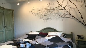 Una cama bien hecha con una colcha a cuadros, dos almohadas con el texto "Steigerhaus", una bandeja con tazas y una tetera, y una obra de arte en la pared que muestra ramas desnudas.