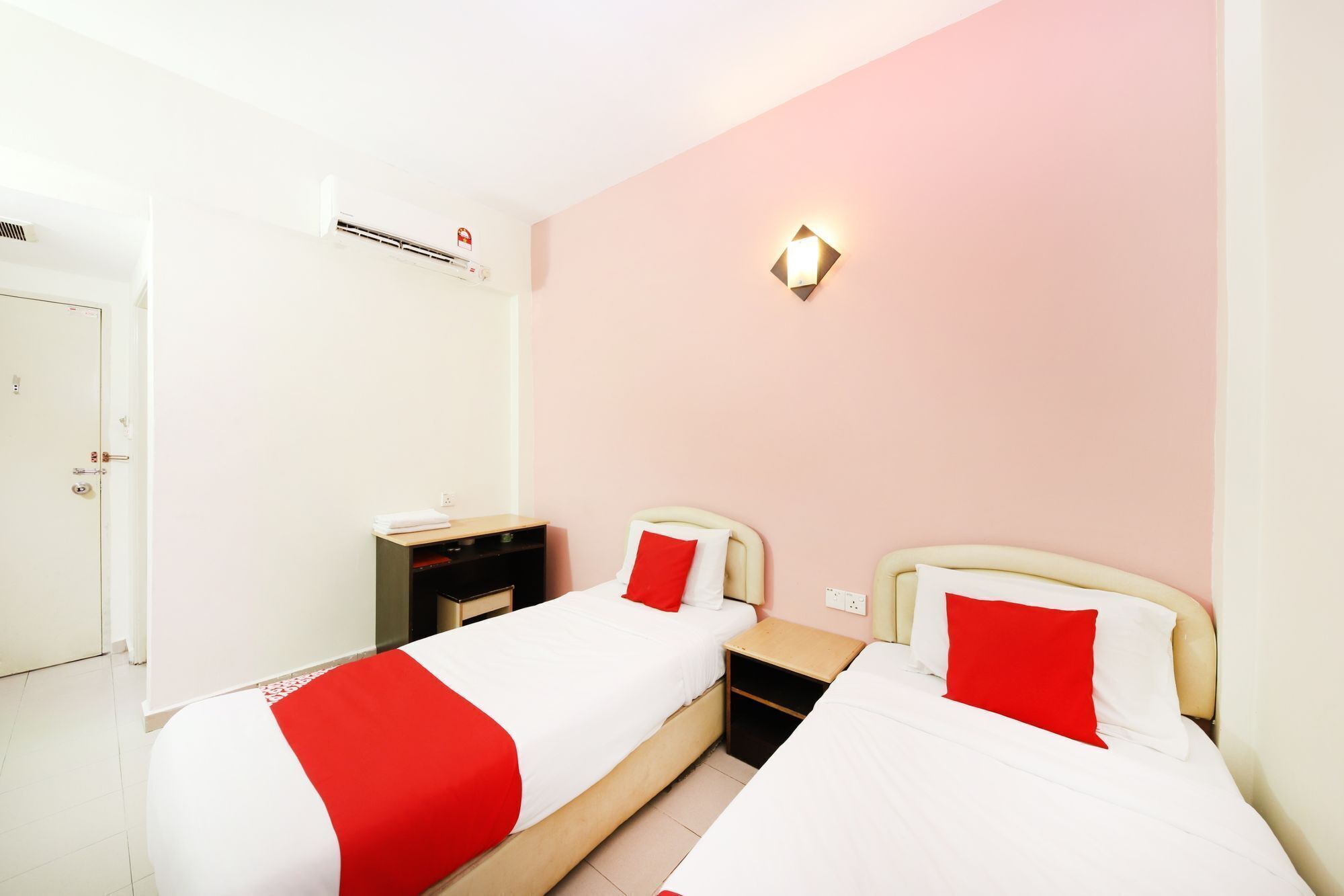 A hotel room with two beds, each with a red and white bedspread, a bedside table, a nightstand, a wardrobe, and a wall-mounted air conditioning unit.
