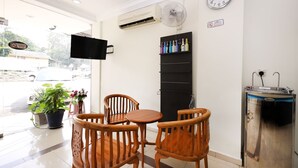 Habitación de hotel con una mesa y sillas de madera, televisor montado en la pared, un reloj, repisa con bebidas embotelladas y una cafetera.