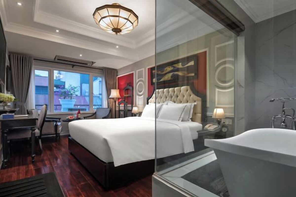 A modern hotel room with a large bed, a desk, and a glass-enclosed bathroom.