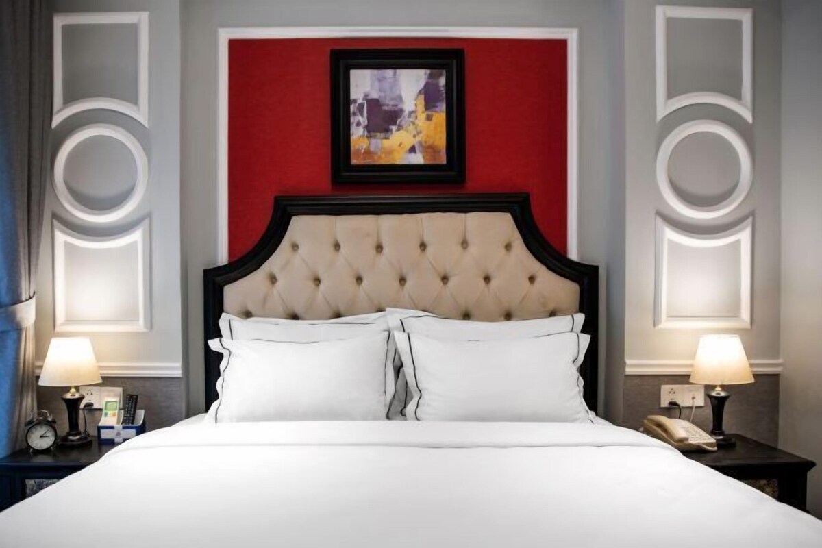A hotel room with a tufted headboard, a red accent wall, a framed abstract painting, and bedside lamps.