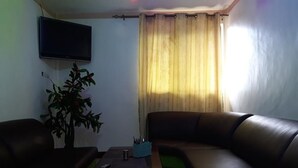 Ein Wohnzimmer mit einer Couch, einem Couchtisch, einem an der Wand montierten Fernseher und einem Fenster mit Vorhängen.