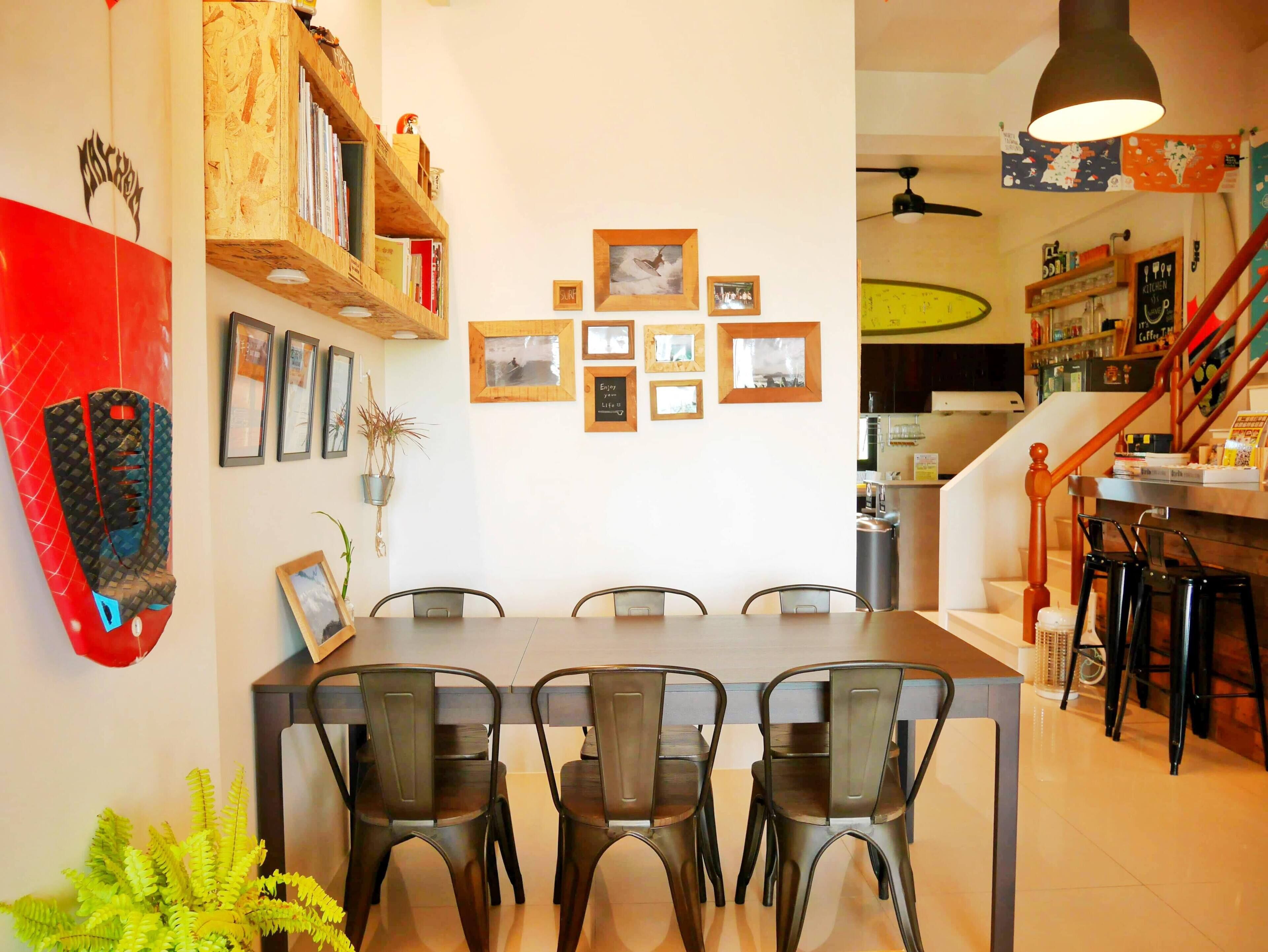 A dining area with a long table and four chairs, a wooden bookshelf, and several framed pictures on the wall.