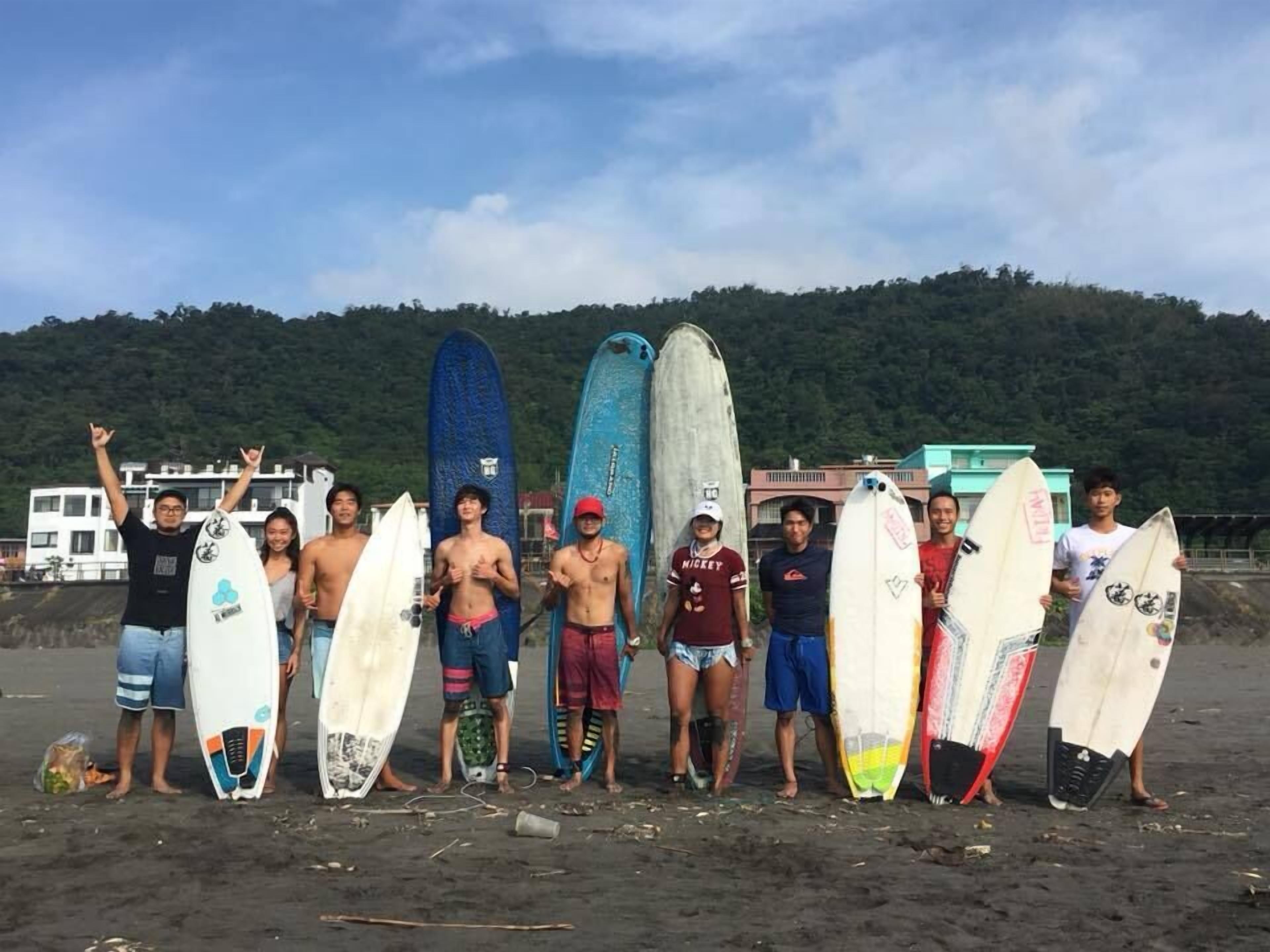 Un groupe de personnes se tient sur une plage, avec des planches de surf, un bâtiment et une colline en arrière-plan.