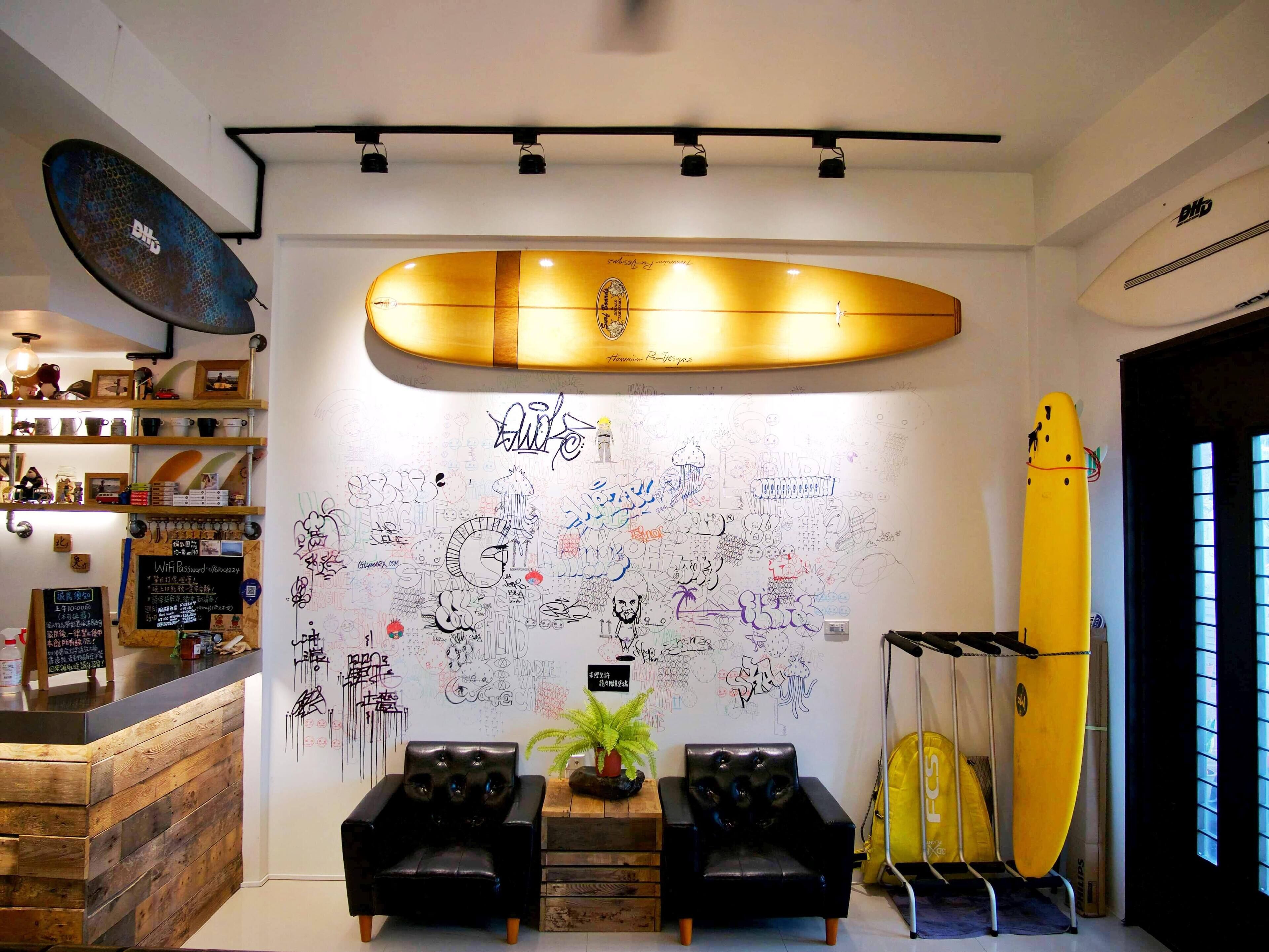A reception area with a surfboard, two black leather chairs, a wall with various drawings, and a wooden counter with shelves.