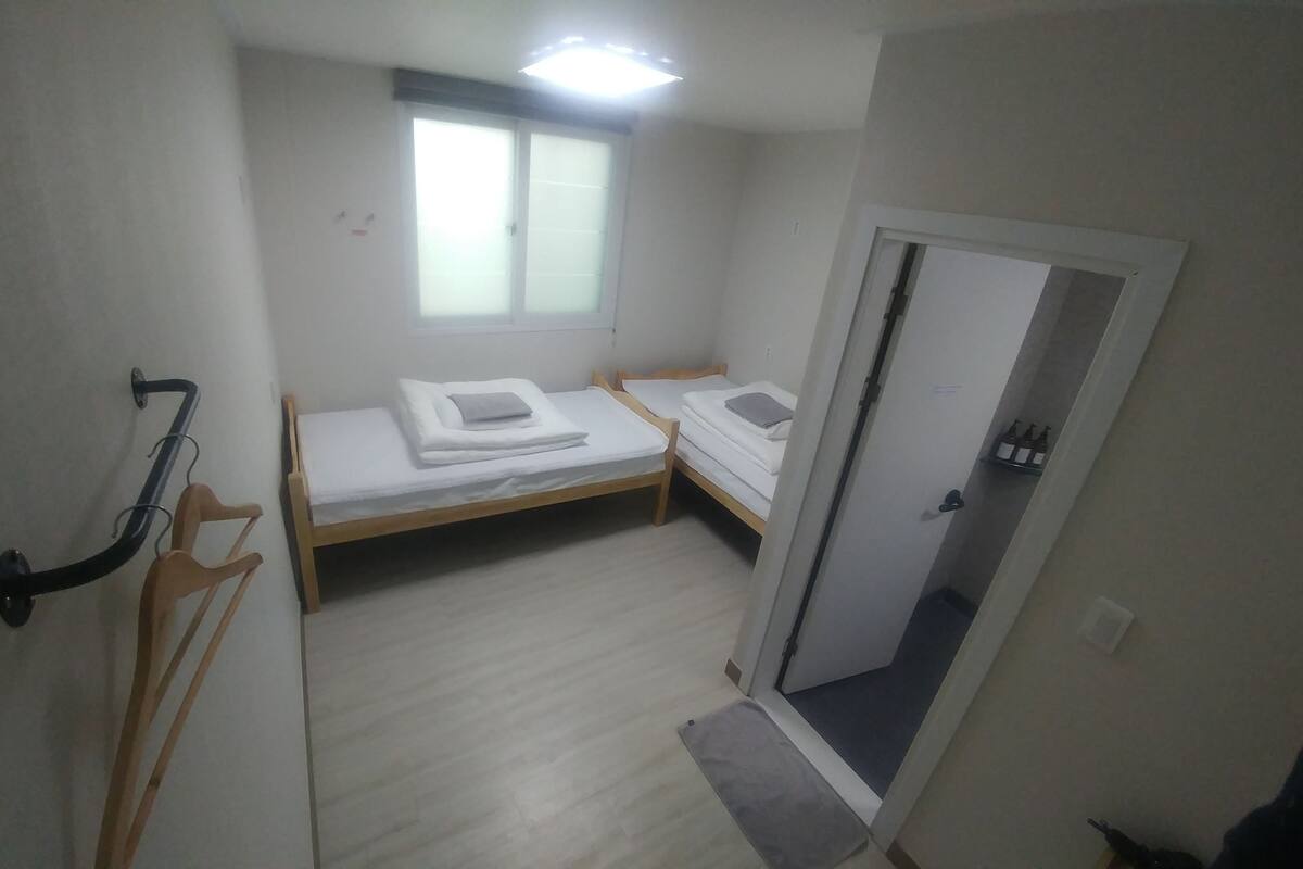 Habitació petita i estreta amb dos llits individuals, una finestra, una porta i una petita estant amb ampolles.