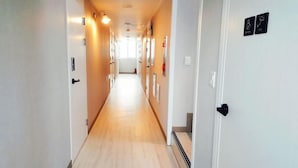 A long, narrow hallway with light-colored walls and wooden flooring. There are doors on both sides, some with black handles and others with round knobs. The hallway is well-lit with wall-mounted lights, and there are several framed pictures on the walls.