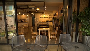 An interior with a dining area featuring round tables and chairs, a kitchen in the background, and a view through a glass door.
