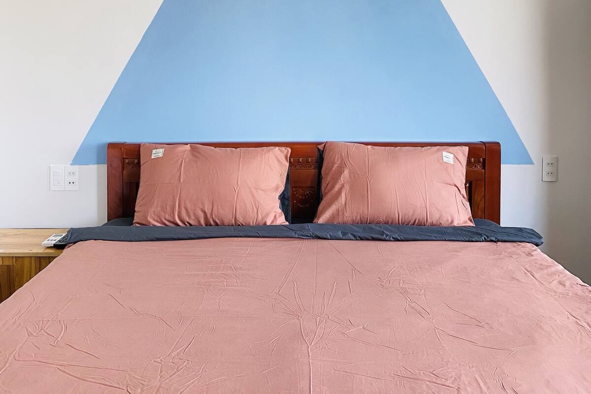 Una cama con una colcha rosa y dos almohadas a juego, un cabecero de madera y un adorno azul en forma de triángulo sobre la cama.