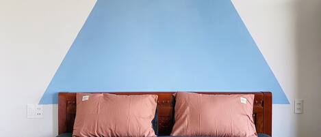 Una cama con una colcha rosa y dos almohadas a juego, un cabecero de madera y un adorno azul en forma de triángulo sobre la cama.