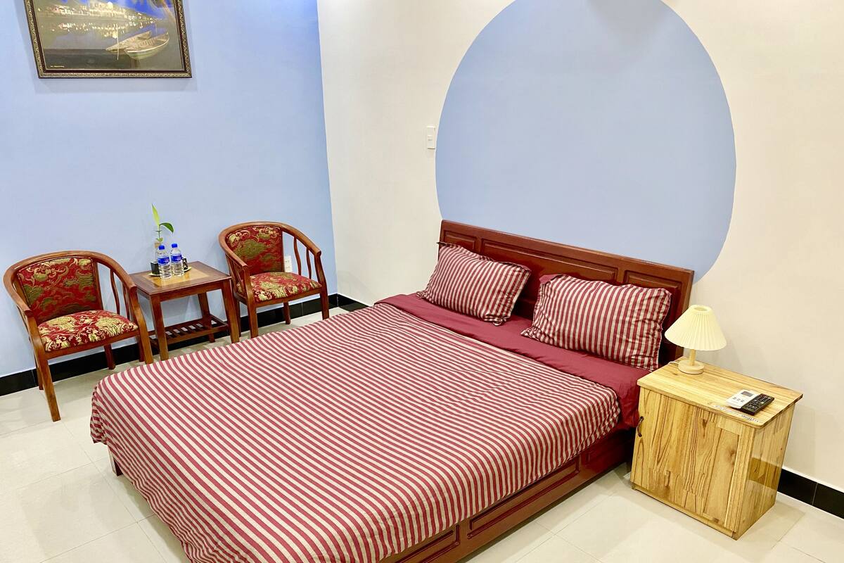Una habitación de hotel ordenada con una cama grande, dos sillas, una mesita y una lámpara de noche.