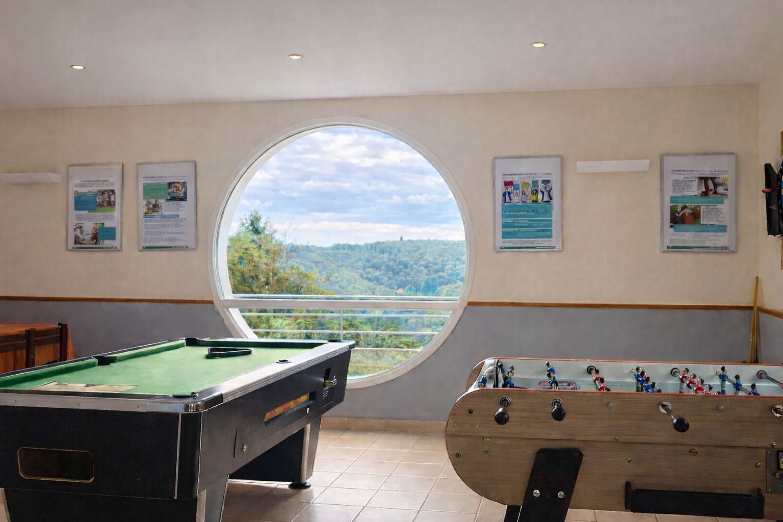 A pool table and foosball table in a room with a large window offering a view of a forested landscape.