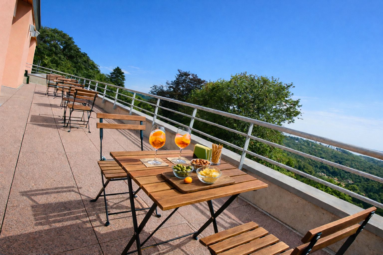 An outdoor terrace with wooden tables and chairs, a railing, and a view of the landscape.