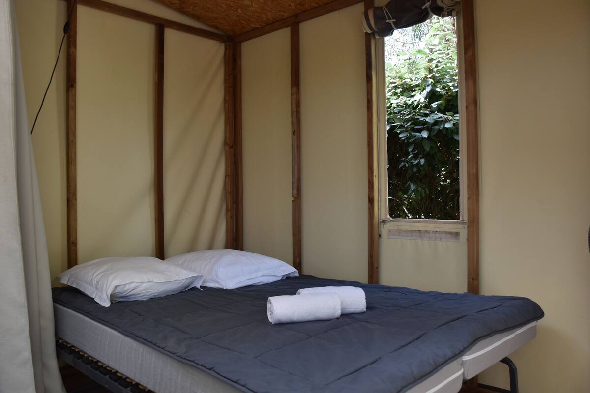 Una cama individual con colchón azul y almohadas blancas, una toalla enrollada y una ventana con vistas a la vegetación.